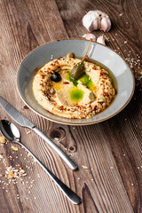 Hummus in a grey bowl seasoned with sunflower and pumpkin seeds, sesame, olives and olive oil. Close-up on a table with cutlery, seeds and garlic by side. Isolated dish on wooden background.