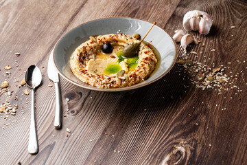 Hummus in a grey bowl seasoned with sunflower and pumpkin seeds, sesame, olives and olive oil. Close-up on a table with cutlery, garlic and seeds by side. Isolated on wooden background. Copy space.
