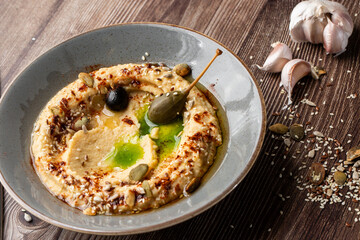 Hummus in a grey bowl seasoned with sunflower and pumpkin seeds, sesame, olives and olive oil. Macro close-up on a table with seeds and garlic by side. Isolated dish on wooden background.