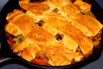 Oven baked dinner in a skillet, strips of crispy biscuit crescent roll lattice across the top. Close up of casserole in a cast iron pan.