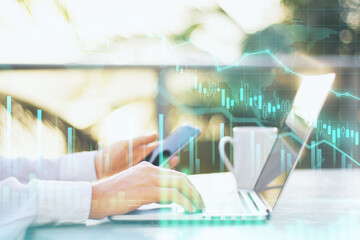 Business crisis concept with businessman hand working on laptop and following stock market news on phone, falling financial chart. Double exposure.
