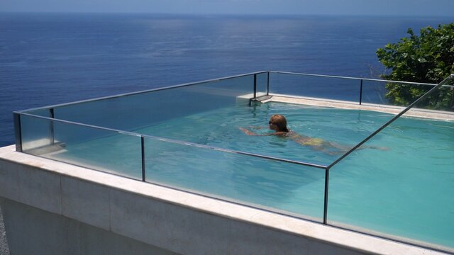Happy middle aged woman swimming in an infinity pool overlooking an ocean. Luxurious resorts concept.