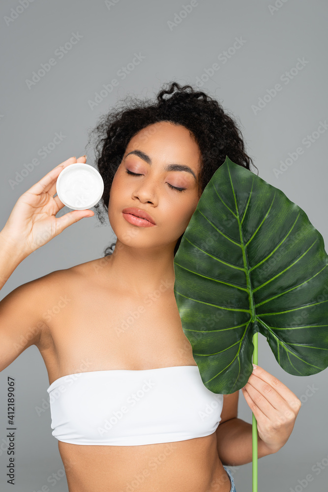 Wall mural African american woman in white top holding cosmetic cream and green leaf isolated on grey