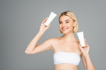 Smiling woman in white top holding tubes with cosmetic lotion isolated on grey