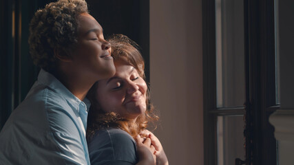Lesbian multiethnic couple hugging standing near window indoors