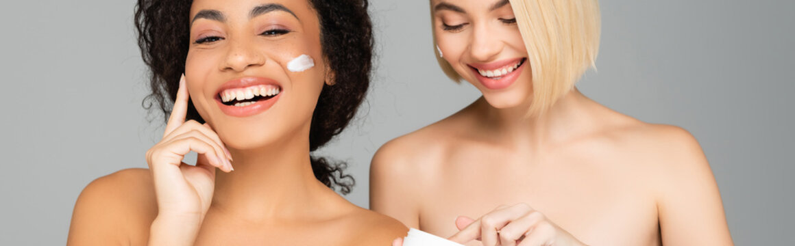 Cheerful multicultural women applying cosmetic cream isolated on grey, banner