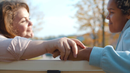 Side view of diverse lesbian couple sitting on bench and holding hands outdoors