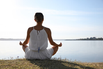 Naklejka na ściany i meble Young woman meditating near river at sunset. Nature healing power