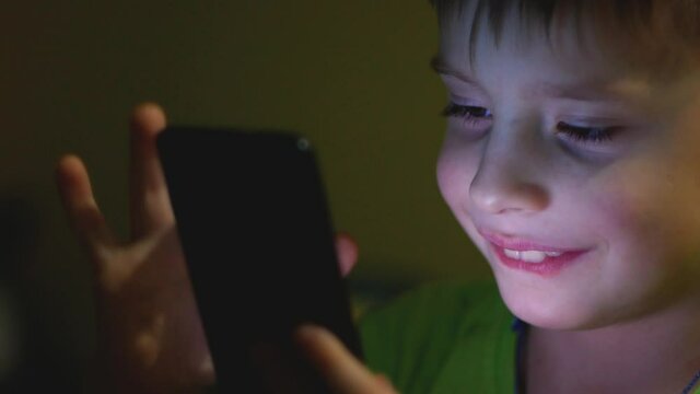 A cute smiling Caucasian boy holds a smartphone in front of his face and surfs the Internet page on a gadget in the dark. The child's face illuminates the light from the gadget screen. Slow motion.