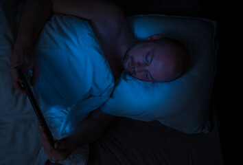 Man lying in bed at night looking at his tablet with harmful blue light illuminating his face.