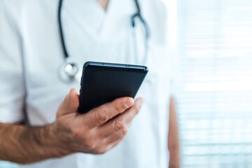 Mature male doctor - nurse wearing face mask, looking at mobile phone next to a hospital window. Covid-19 and medicine concept