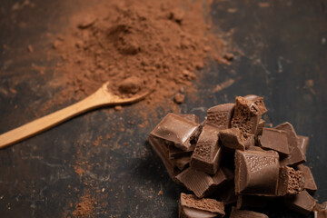 Different pieces of dark chocolate and cocoa powder on a dark surface. Space for text