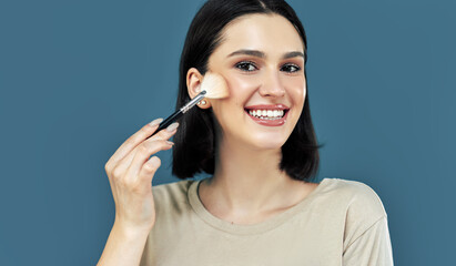 Candid portrait of a happy brunette young woman looking to the camera and making makeup with a brush herself.