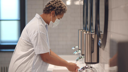 African woman wearing protective face mask washing hands in public bathroom