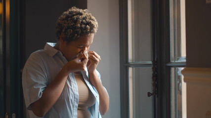 Fototapeta premium African female standing near window and sneezing in paper tissue having pollen allergy