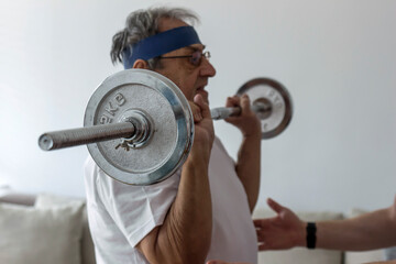 Fototapeta na wymiar Cropped shot of a senior man working out at home. Elder man using dumbbells in order to maintain his physical activity at home in the living room. Photo of a old man is doing sports, recovery concept.
