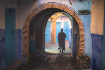 A Moroccoan man in a traditional djellaba walks under an arch in a colourful street scene in the old town medina of Chefchaouen, Morocco with its blue buildings.