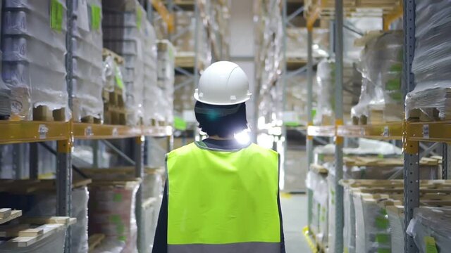 Female warehouse worker wearing hard hat checks walks in storehouse, storeroom with rows of shelves with cardboard boxes. architect walking through logistics of factory. woman in storage with goods