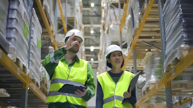Young woman and man are talking and walking at warehouse during working day. Front view of american employees discussing work and going in storehouse, guy holding tablet in hand. People wearing