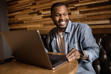 Happy African American uses laptop, looks to the side, chatting with someone in the office, reads good news by email. Young business man is discussing a work plan. Work and business concept.