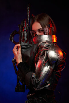 Beautiful cyber woman holding rifle and hiding her face in dark background. Dressed in black jacket martial woman in cyberpunk style looks at camera.