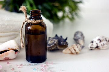 Dark glass cosmetic bottle surrounded by seashells, towels, greenery on a white table, copy space, mock up. Natural organic home cosmetics wellness concept