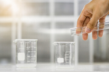 Female hand holding glass tube above beaker on white table on science laboratory background.