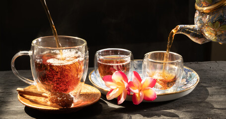 Pouring freshly brewed  tea glass cup with Plumeria flower,cinnamon sugar stick and traditional kettle on dark wood table,top view.