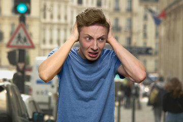 Teenager's having nervous breakdown. Crying european boy is closing his ears with his hands. Young boy wants silence. Noised city on the background.
