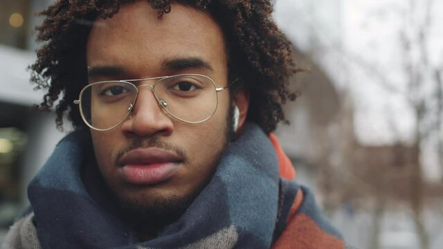 Close up portrait shot of young handsome Afro-American man in glasses, wireless earphones and warm scarf standing on winter day and looking at camera