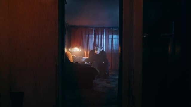 Eerie scene of a scary, masked stranger sitting in a dark living room, petting a cat. Mysterious man in an animal mask, goat costume. Devil waiting in the night, dark, unsettling scene.