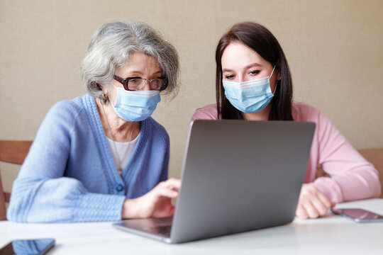 An elderly woman and a young woman wearing masks on her face use a laptop at home amid coronavirus - A volunteer helping an elderly person to use modern technologies