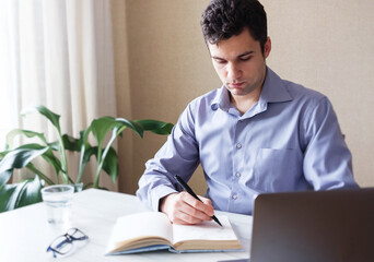Fototapeta premium Millennial learns online and makes notes in his notebook - Student learns through video communication - A business person conducts business from his home