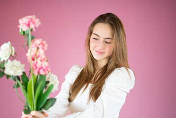 Portrait of a beautiful young girl in white shirt holding big bouquet of flowers isolated over pink background