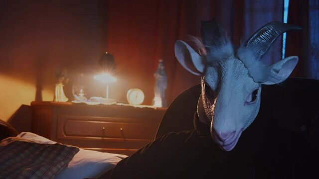 Eerie scene of a scary, masked stranger sitting in a dark living room, petting a cat. Mysterious man in an animal mask, goat costume. Devil waiting in the night, dark, unsettling scene.