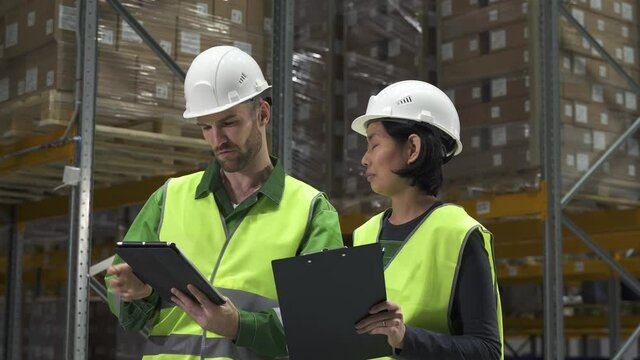 man and asian woman two worker in hard hat with clipboard talking working process. warehouse manager looking at tablet checking inspecting rows with goods. concept logistics, racks of boxes.