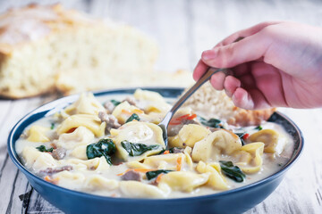 Hand with Tortellini Soup with Italian sausage, spinach and carrots. Served with homemade artisan bread over a white wood wooden table. Selective focus on soup in spoon with blurred background.