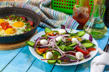 Fried eggs in a plate with sausages, cherry tomatoes and bell pepper for breakfast, and a plate of Greek salad, on a blue wooden background