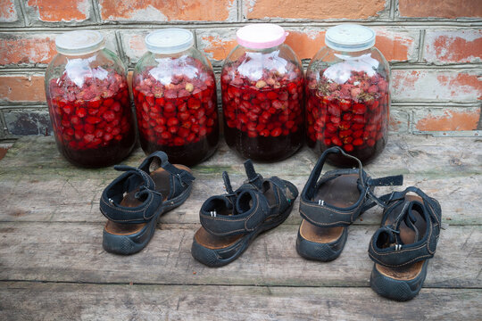 A country courtyard, against the wall of a red brick house, there are four three-liter jars of strawberry compote. Childrens shoes are standing in front of the banks.