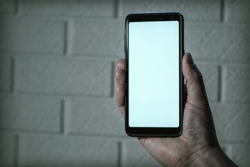 Smartphone mockup. A hand holds a black phone with a white blank screen against a white brick wall background.