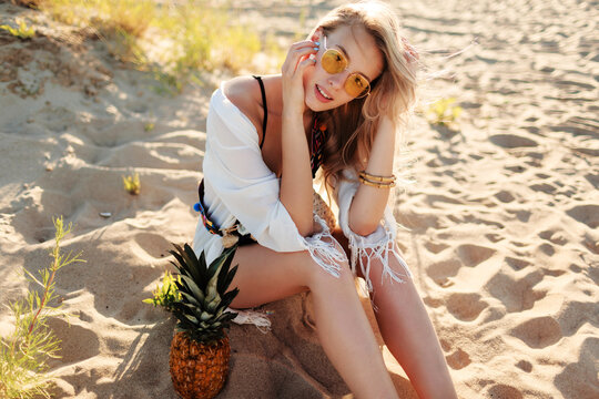 Lifestyle outdoor picture of laughing pretty woman with juicy pineapple relaxing on sunny beach . Trendy summer outfit