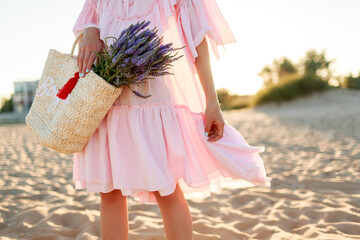 Romantic blonde  female in cute pink  dress dancing and having fu on  the beach. Holding straw bag...