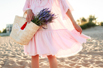 Romantic blonde  female in cute pink  dress dancing and having fu on  the beach. Holding straw bag...