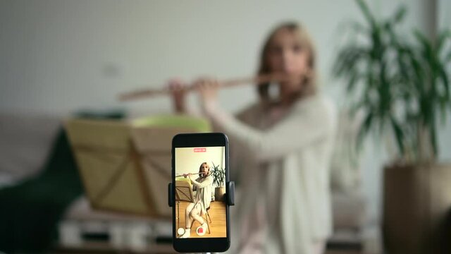 Woman play flute, wind instrument and record video with phone for social media. Solo musician film vlog on smartphone. Mature middle aged flutist practice with sheet music. Concept internet tutorial