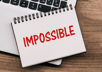 A notebook with the text IMPOSSIBLE lies on a laptop standing on a wooden table.