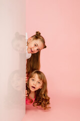 Happy kids, girls isolated on coral pink studio background. Look happy, cheerful. Copyspace for ad. Childhood, education, emotions, business, facial expression concept. Peeking out from behind the