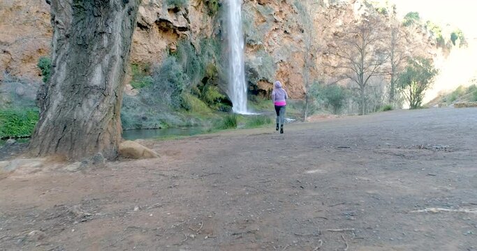 Mujer con ch&aacute;ndal corriendo en paraje con cascada y dron volando tras ella