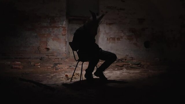 Possessed maniac tied to a chair. Broken light over a tortured person in animal mask. Eerie, frightening scene, scary basement.