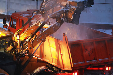 An excavator loads snow into the back of a dump truck at night. The work of road and municipal services for the removal of snow in winter.