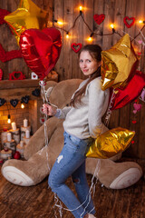 happy girl in beautiful backdrop for Valentine's Day photo shoot with huge teddy bear, wooden fireplace with candles, with a bunch of balloons and flowers, and tree with red and pink hearts.